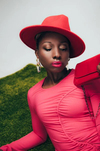 Atractiva mujer afroamericana en vestido rosa y sombrero posando con bolsa en hierba verde - foto de stock