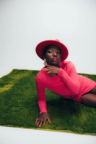 Seductora mujer afroamericana en vestido rosa y sombrero posando sobre hierba verde - foto de stock