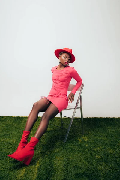Модна афроамериканка в рожевій сукні і капелюсі позує на стільці на зеленій траві — стокове фото