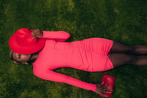 Vista superior de la elegante mujer afroamericana en vestido rosa posando con sombrero y bolso sobre hierba verde - foto de stock