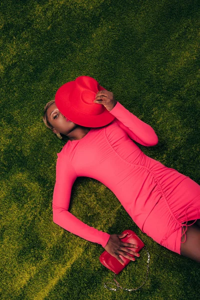 Vista superior da bela mulher americana africana em vestido rosa posando com chapéu e saco na grama verde — Fotografia de Stock