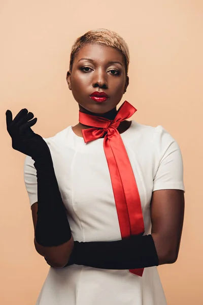 Joven mujer afroamericana posando en vestido blanco, bufanda roja y guantes negros aislados en beige - foto de stock