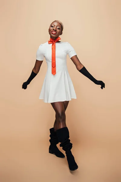 Niña afroamericana sonriente posando en vestido blanco, bufanda roja y guantes negros en beige - foto de stock