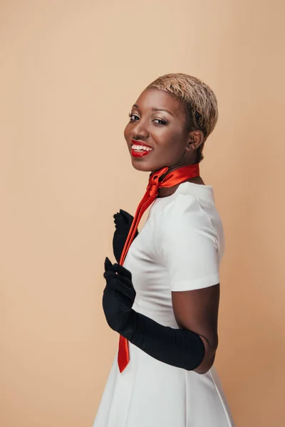 Mulher americana africana alegre posando em vestido branco, cachecol vermelho e luvas pretas isoladas no bege — Fotografia de Stock