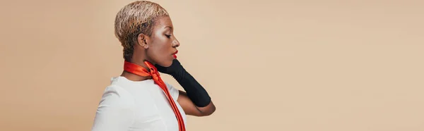 Елегантна афроамериканка з коротким волоссям позує в білій сукні, червоний шарф і чорні рукавички ізольовані на бежевому — стокове фото