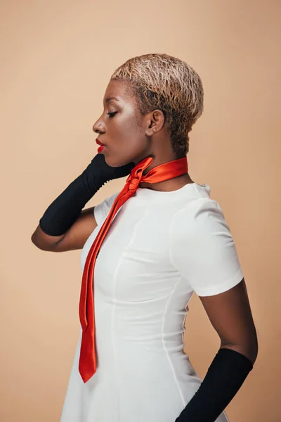 Afro-américaine fille aux cheveux courts posant en robe blanche, écharpe rouge et gants noirs isolés sur beige — Photo de stock