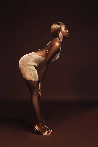 Вид сбоку чувственной африканской женщины с короткими волосами, позирующей в гламурном платье коричневого цвета — стоковое фото