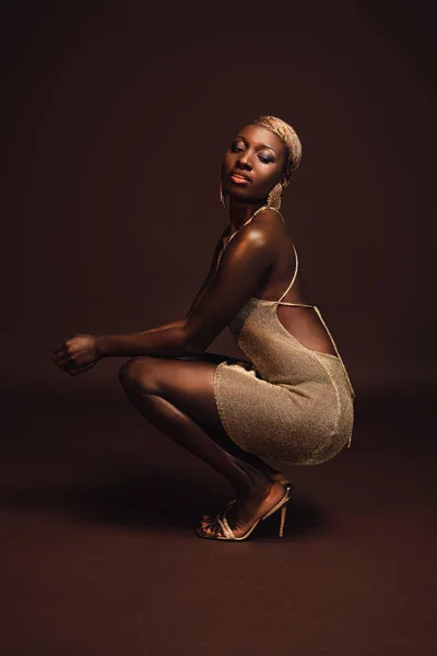 Стильная африканская американка с короткими волосами, позирующая в гламурном платье на коричневом — стоковое фото