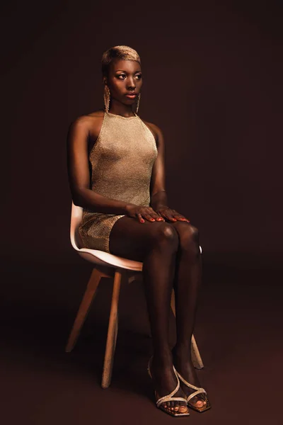 Красивая стильная африканская американка с короткими волосами, сидящая на стуле на коричневом — стоковое фото