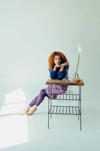 Stylish redhead girl posing at table with calla flower and vases on grey — Stock Photo