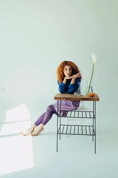 Chica pelirroja elegante posando en la mesa con flor de calla y jarrones en gris - foto de stock