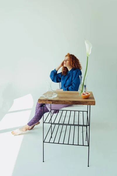 Chica pelirroja de moda posando en la mesa con flor de calla y jarrones en gris - foto de stock