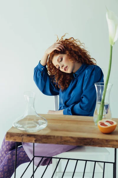 Beautiful fashionable redhead girl posing at table with calla flower and vases on grey — Stock Photo