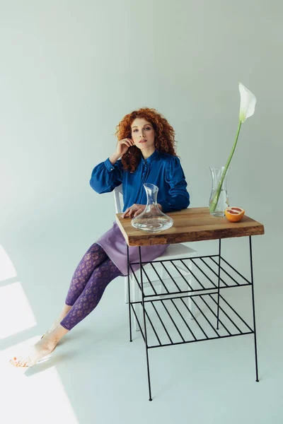 Beautiful fashionable redhead girl posing at table with calla flower and vases on grey — Stock Photo