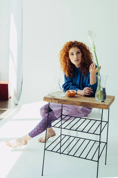 Beautiful fashionable redhead girl posing at table with calla flower and vases on white — Stock Photo