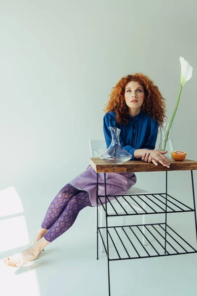 Attractive fashionable redhead girl posing at table with calla flower and vases on grey — Stock Photo