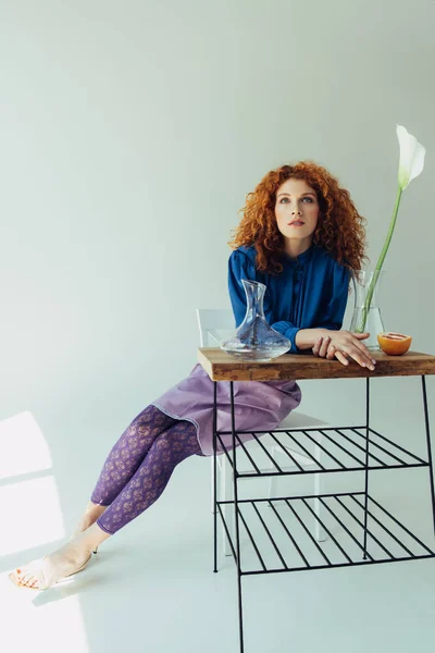 Attrayant rousse fille à la mode posant à la table avec fleur de calla et vases sur gris — Photo de stock