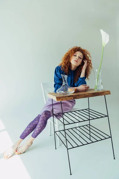 Beautiful trendy redhead girl posing at table with calla flower and vases on grey — Stock Photo