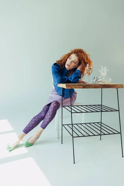 Beautiful stylish redhead girl posing at table with glass and vase on grey — Stock Photo