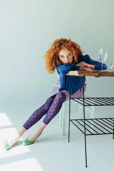 Beautiful stylish redhead girl posing at table with glass on grey — Stock Photo
