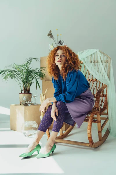 Chica pelirroja elegante sentado en silla de mimbre cerca de cajas de madera y flores secas en gris - foto de stock