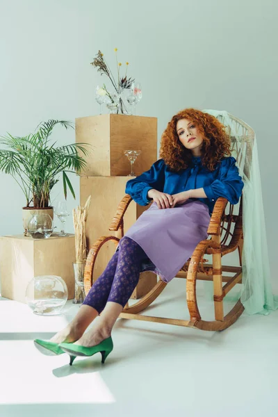 Chica pelirroja elegante sentado en silla de mimbre cerca de cajas de madera, vasos y flores secas en gris - foto de stock