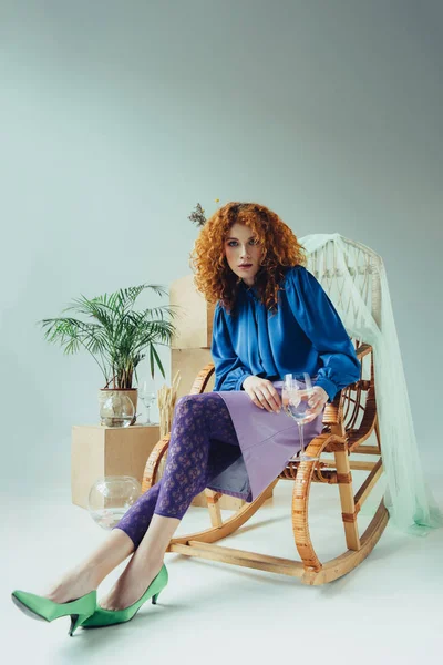 Stylish redhead girl sitting in wicker chair near wooden boxes and plant on grey — Stock Photo