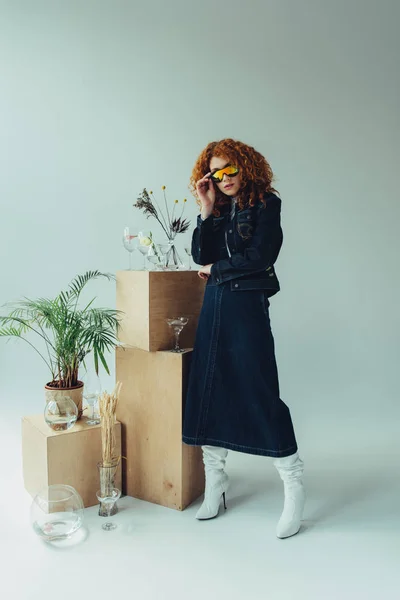 Стильная рыжая девушка в солнечных очках позирует возле коробок, стаканов и растений на сером — стоковое фото