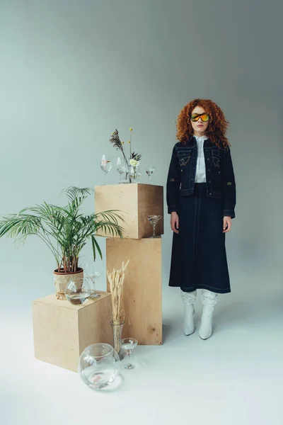 Fashionable redhead girl posing near wooden boxes, glasses and plants on grey — Stock Photo