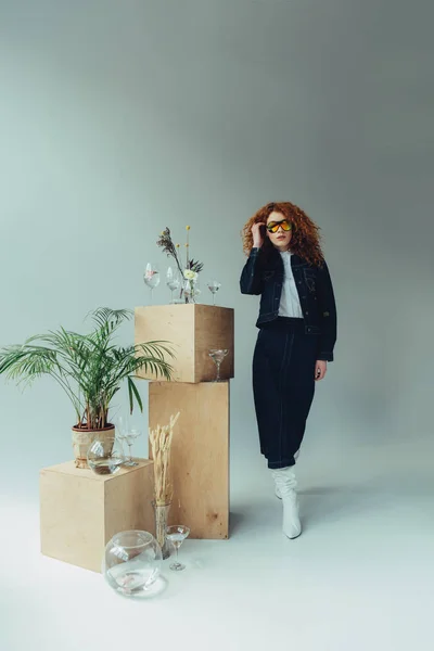 Fashionable redhead girl posing near wooden boxes, glasses and plants on grey — Stock Photo