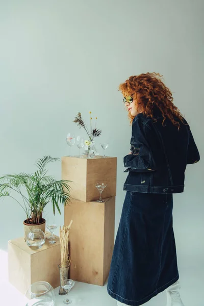 Menina ruiva elegante em óculos de sol perto de caixas de madeira, óculos e plantas em cinza — Fotografia de Stock