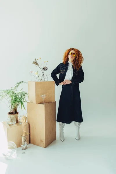 Élégant rousse fille dans des lunettes de soleil près de boîtes en bois, des lunettes et des plantes sur gris — Photo de stock