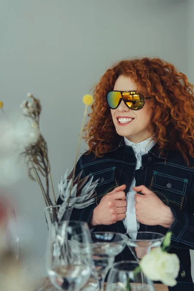 Sonriente mujer pelirroja con estilo en gafas de sol posando cerca de gafas y flores secas aisladas en gris - foto de stock