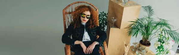 Panoramic shot of stylish redhead girl in wicker chair near wooden boxes, glasses and plants on grey — Stock Photo