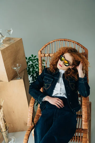 Chica pelirroja con estilo en silla de mimbre cerca de cajas de madera y gafas aisladas en gris - foto de stock