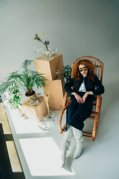 Vue grand angle de fille rousse élégante dans une chaise en osier près des boîtes en bois, des lunettes et des plantes — Photo de stock