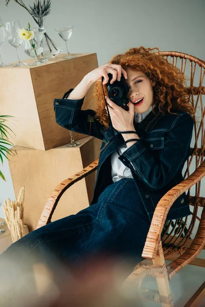 Stylish redhead woman in wicker chair taking photo with film camera on grey — Stock Photo