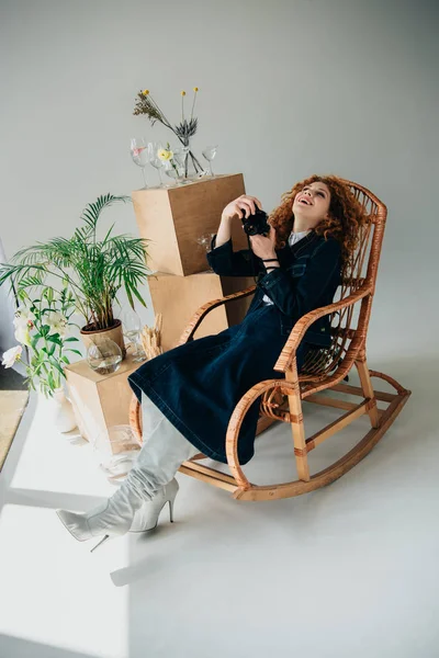 Стильная смеющаяся девушка в плетеном кресле с кинокамерой рядом с деревянными коробками, стаканами и растениями на сером — стоковое фото