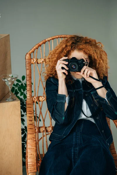 Stylish redhead woman in wicker chair taking photo with film camera on grey — Stock Photo