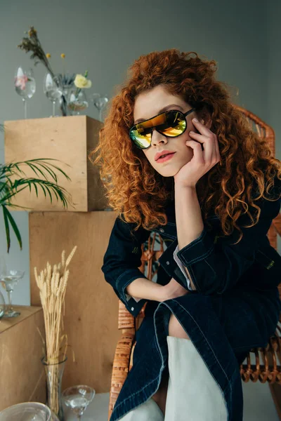 Stylish redhead girl in sunglasses in wicker chair near wooden boxes, glasses and plants on grey — Stock Photo