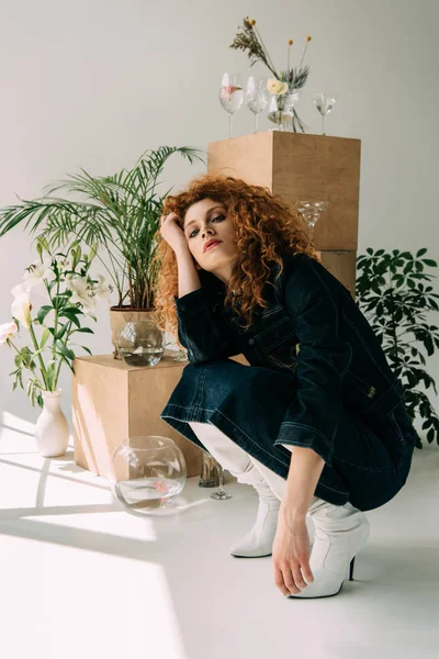 Trendy redhead girl sitting and posing near wooden boxes, glasses and plants on grey — Stock Photo