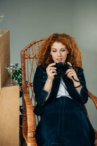 Belle femme rousse élégante dans une chaise en osier prenant des photos avec caméra de film sur gris — Photo de stock