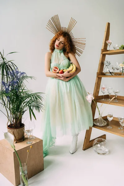 Élégant rousse fille avec accessoire sur la tête et les fruits près de l'échelle avec des fleurs et des lunettes — Photo de stock