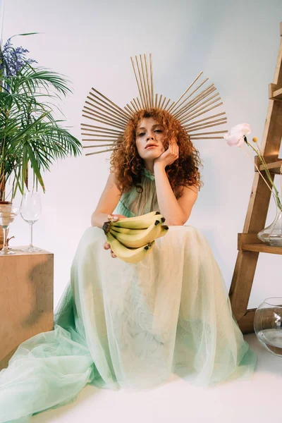 Stylish redhead woman with accessory on head posing with bananas and propping head — Stock Photo