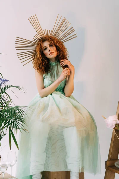 Fashionable redhead girl with accessory on head in dress posing near plant — Stock Photo