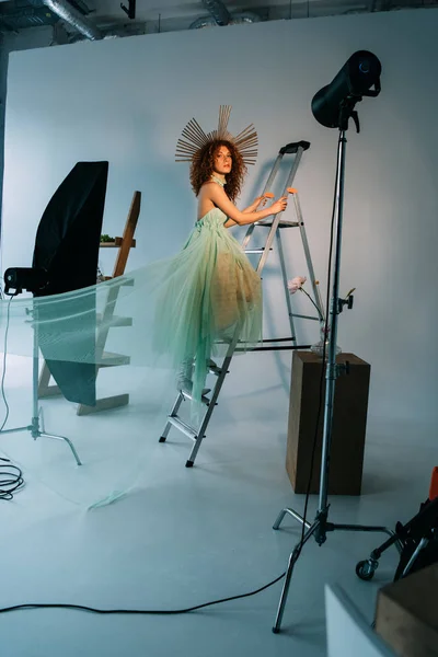 Beautiful stylish girl with accessory on head posing on ladder near photographic lightning — Stock Photo