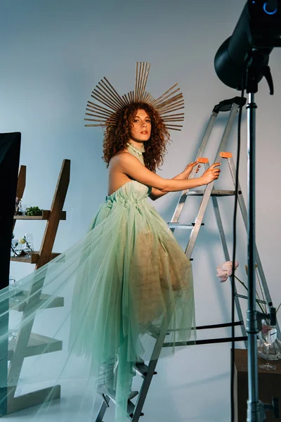 Beautiful stylish girl with accessory on head posing on ladder near photographic lightning — Stock Photo