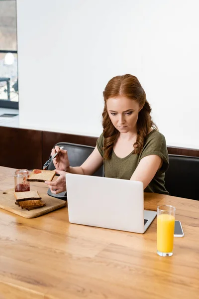 Выборочный фокус женщины, смотрящей на ноутбук, держа тост с джемом за столом — стоковое фото