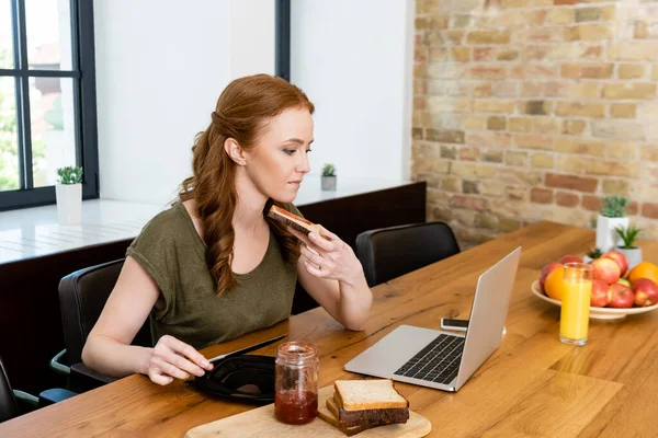 Frau hält Toast und Messer neben digitalen Geräten und Orangensaft auf Tisch — Stockfoto