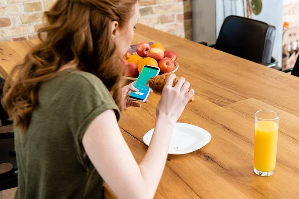 КИЕВ, УКРАИНА - 8 мая 2020 года: вид сбоку женщины, использующей смартфон с приложением twitter во время еды круассана за столом — стоковое фото